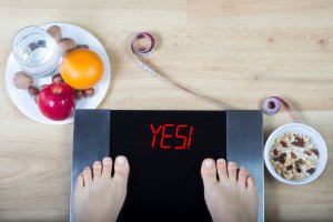 Person standing on a digital scale showing weight loss, surrounded by healthy foods, water, and a measuring tape, representing metabolic balance results